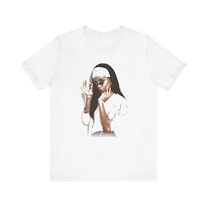 May include: White t-shirt featuring a graphic print of a woman wearing a white bandana, sunglasses, and a white jacket. The woman is posing with her hands in the air.