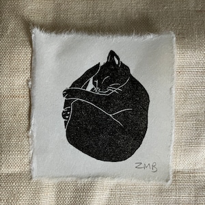 Könnte beinhalten: Schwarz-weiß-Illustration einer Katze, die sich zusammengerollt schlafend. Das Bild ist auf einem weißen Stoff mit rauer Textur gedruckt. Die Initialen des Künstlers "ZMB" sind unten rechts signiert.