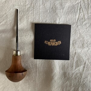 May include: A wood-handled metal carving tool with a rounded grip and a thin, pointed blade, positioned next to a square black card featuring a gold-colored Claddagh symbol printed in the center, depicting two hands holding a crowned heart.