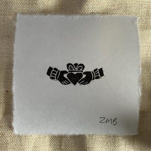 May include: A black and white print of a Claddagh ring design on a square piece of paper. The Claddagh symbol features two hands clasping a heart, topped with a crown. The initials "ZMB" are in the lower right corner.