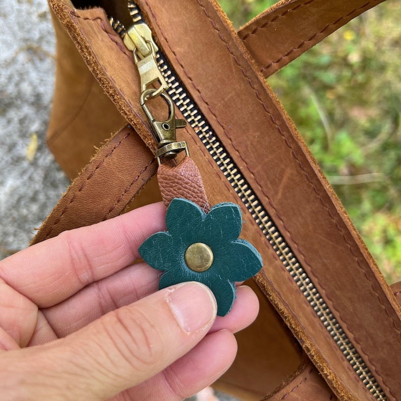Hand holding a green leather flower charm near a brown leather bag with a gold zipper.