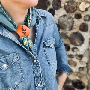 May include: A person wearing a blue denim shirt with a green and white patterned bandana tied around their neck. The bandana is secured with a leather bolo tie featuring a snake design.