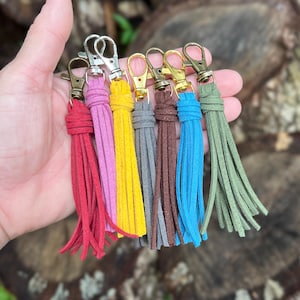 May include: A collection of seven suede tassels in red, pink, yellow, gray, brown, blue, and olive green. Each tassel is attached to a metal lobster claw clasp, featuring a variety of metallic finishes including silver, gold, and antique bronze tones.
