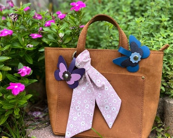 May include: A brown leather tote bag with a floral print scarf and two leather butterfly charms. One butterfly charm is purple and the other is blue.