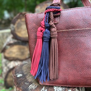 May include: A dark brown pebbled leather handbag featuring a trio of suede tassels in red, navy blue, and brown. The tassels are attached to the bag handle with braided cords. The bag is embossed with the text PORTLAND LEATHER GOODS.