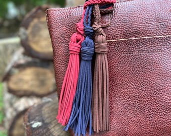 May include: A dark brown pebbled leather handbag featuring a trio of suede tassels in red, navy blue, and brown. The tassels are attached to the bag handle with braided cords. The bag is embossed with the text PORTLAND LEATHER GOODS.