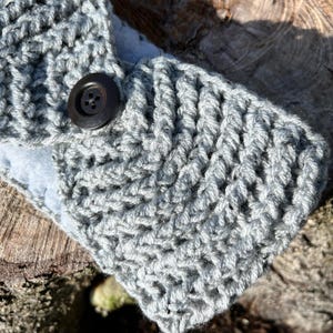 May include: Close-up of a gray crocheted headband with a large, dark brown button closure. The headband features a textured stitch pattern and a soft, light blue lining. The item is handmade.