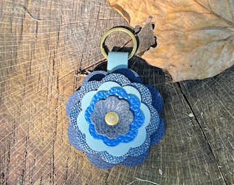 May include: A layered flower-shaped keychain featuring multiple shades of blue and grey textured material. The petals are stacked in a circular pattern with a central gold-toned metal rivet and a gold-toned metal split ring at the top.