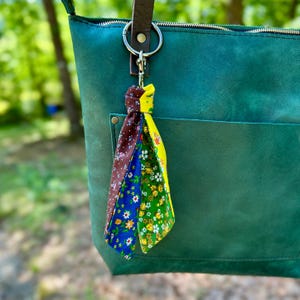 May include: A colorful fabric tassel keychain with a gold-tone clasp hangs from a teal green leather tote bag. The tassel features brown, blue, and yellow floral patterned fabric strips. The bag has a zipper closure and a front pocket.