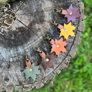 May include: Five maple leaf-shaped keychains made of leather in shades of burgundy, yellow, orange, brown, and sage green. Each keychain features a textured brown leather strap, a brass-toned metal lobster clasp, and a central brass-toned rivet.