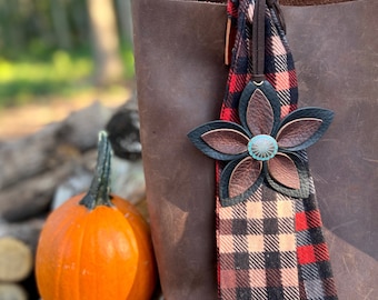 May include: A brown leather tote bag with a decorative leather flower charm. The flower has brown and black petals with a turquoise center. A red and black plaid scarf is attached to the bag.