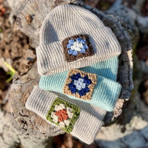 May include: Three ribbed knit beanies stacked vertically. The top beanie is beige with a brown and blue crochet granny square patch. The middle beanie is light blue with a brown and blue crochet patch. The bottom beanie is beige with a green and orange patch.
