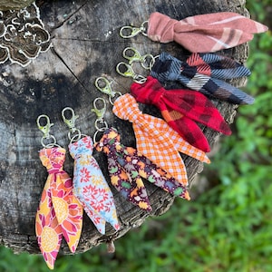 May include: A collection of seven fabric keychains featuring various patterns including floral, plaid, and leaf prints. Each keychain is constructed from knotted fabric strips attached to a silver-toned metal lobster clasp for easy attachment to bags or keys.