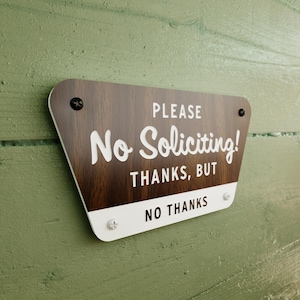 May include: A wooden sign with white text that reads "Please No Soliciting! Thanks, But No Thanks". The sign has a brown woodgrain pattern and is mounted on a green wall.