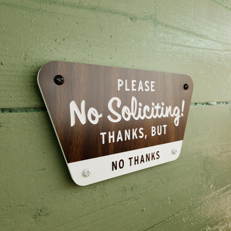 May include: A wooden sign with white text that reads "Please No Soliciting! Thanks, But No Thanks". The sign has a brown woodgrain pattern and is mounted on a green wall.