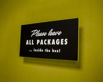 May include: A black sign with white text that reads "Please leave ALL PACKAGES... inside the box!" The sign is mounted on a yellow wall.