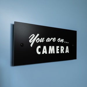 May include: Black sign with white text that reads "You are on... CAMERA".