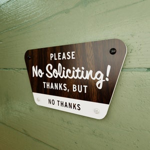 May include: A brown wooden sign with white text that reads "PLEASE No Soliciting! THANKS, BUT NO THANKS". The sign has rounded corners and is attached to a green wall with screws.