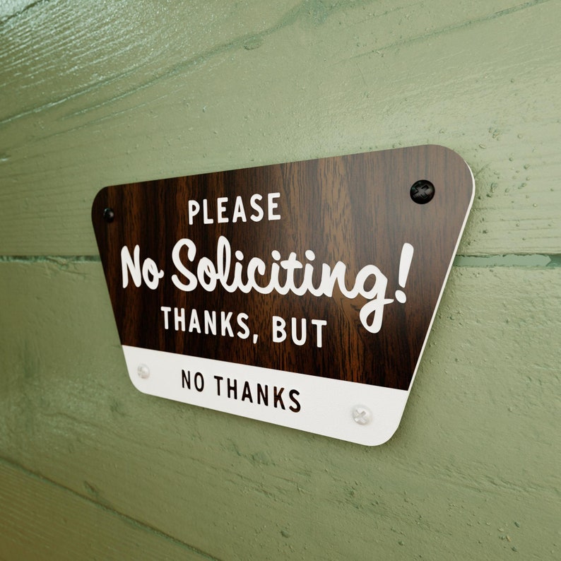 May include: A brown wooden sign with white text that reads "PLEASE No Soliciting! THANKS, BUT NO THANKS". The sign has rounded corners and is attached to a green wall with screws.