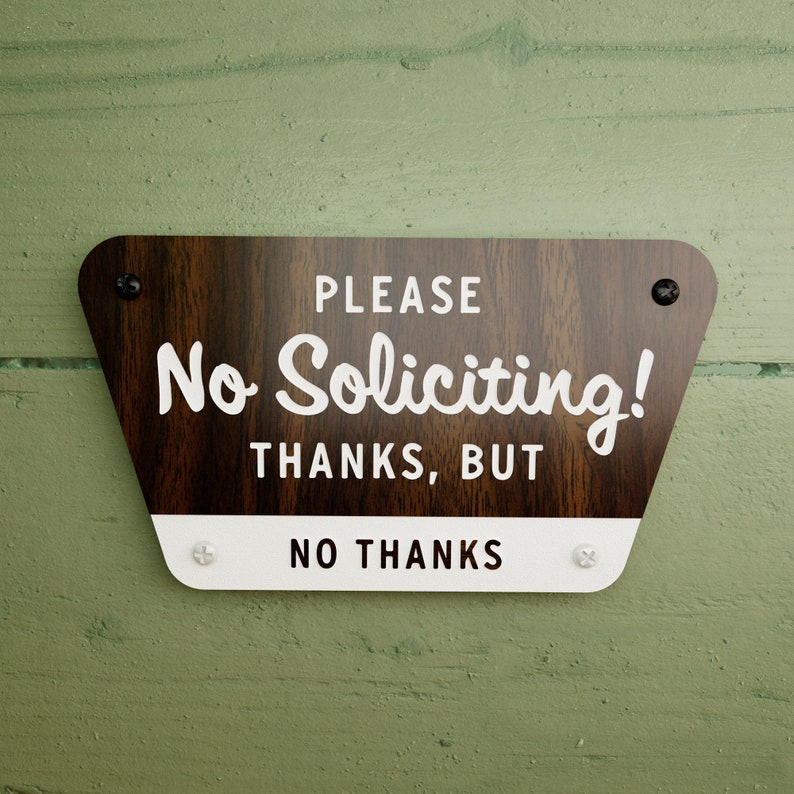 May include: A brown wooden sign with rounded edges and white text that reads "PLEASE No Soliciting! THANKS, BUT NO THANKS". The sign has two black screws in the top corners and two white screws in the bottom corners.