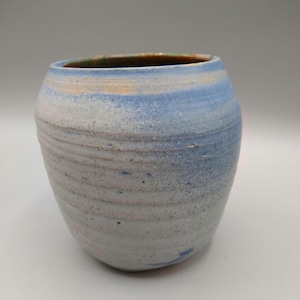 May include: A handmade ceramic vase with a rounded shape and a wide opening. The vase features a textured surface with horizontal bands of color, including light blue, white, and tan. The rim has a green and brown hue.