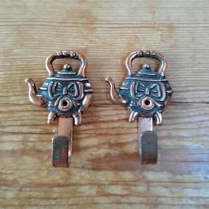 May include: Two decorative copper-colored metal wall hooks. Each hook is shaped like a teapot with a dark teal accent and a bow design. The hooks are mounted on a wooden surface.