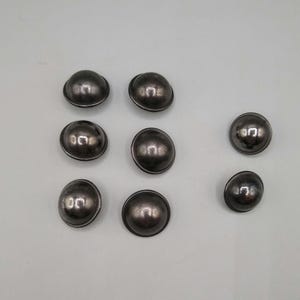 May include: A set of seven circular, dome-shaped buttons with a dark, metallic gunmetal finish. Each button features a smooth, reflective surface with a raised center and a thin, defined outer rim, arranged in a loose cluster on a neutral background.
