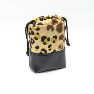 May include: A small drawstring bag with a black leather base and a tan fabric top. The fabric top features a leopard print pattern in shades of brown, tan, and black. The bag has a black drawstring closure.