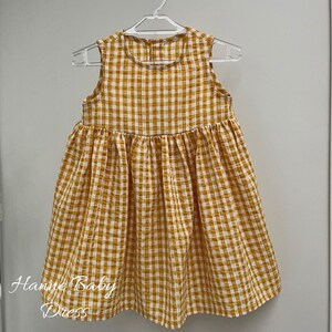 May include: A sleeveless, yellow and white checkered baby dress hanging on a white hanger. The dress has a gathered skirt and the text "Hanne Baby Dress" is visible on the lower left side.