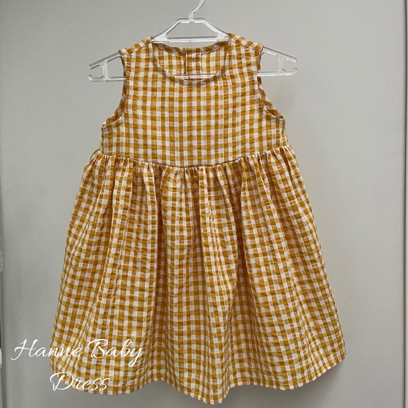 May include: A sleeveless, yellow and white checkered baby dress hanging on a white hanger. The dress has a gathered skirt and the text "Hanne Baby Dress" is visible on the lower left side.