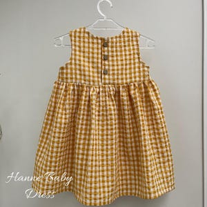May include: A sleeveless baby dress in a yellow and white gingham pattern, hanging on a clear plastic hanger. The dress has three brown buttons on the back and the text "Hanne Baby Dress" in cursive at the bottom.