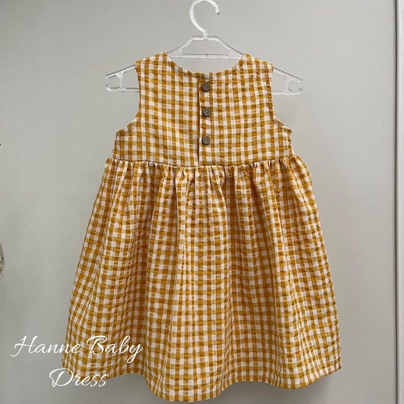 May include: A sleeveless baby dress in a yellow and white gingham pattern, hanging on a clear plastic hanger. The dress has three brown buttons on the back and the text "Hanne Baby Dress" in cursive at the bottom.