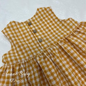 May include: A yellow and white gingham baby dress with three wooden buttons down the back. The dress has a gathered skirt and the text "Hanne Baby Dress" is printed on the front.
