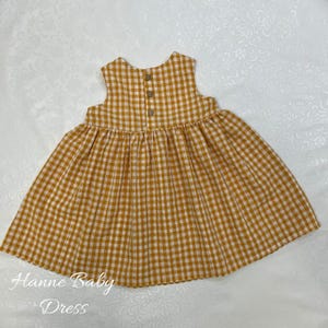 May include: A sleeveless baby dress in a yellow and white gingham pattern. The dress has a gathered skirt and three wooden buttons on the back. The text "Hanne Baby Dress" is printed on the bottom left.
