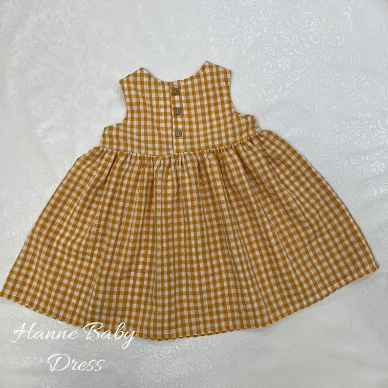 May include: A sleeveless baby dress in a yellow and white gingham pattern. The dress has a gathered skirt and three wooden buttons on the back. The text "Hanne Baby Dress" is printed on the bottom left.