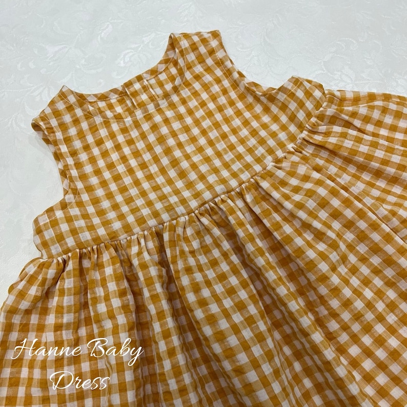 May include: A sleeveless baby dress in a yellow and white gingham pattern. The dress features a gathered waist and a high neckline. The text "Hanne Baby Dress" is printed in cursive at the bottom left of the image.