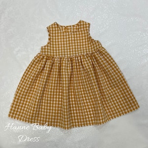 May include: A sleeveless, mustard yellow and white gingham baby dress. The dress has a gathered skirt and a round neckline. The text "Hanne Baby Dress" is printed in cursive at the bottom of the image.