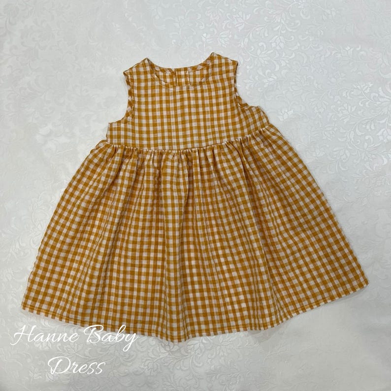 May include: A sleeveless, mustard yellow and white gingham baby dress. The dress has a gathered skirt and a round neckline. The text "Hanne Baby Dress" is printed in cursive at the bottom of the image.