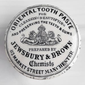 May include: A white, circular container with black text, advertising "Oriental Tooth Paste." The text includes "Cleansing, Beautifying, and Preserving the Teeth & Gums." Features a crest with lions and a crown. Prepared by Jewsbury & Brown, Chemists.