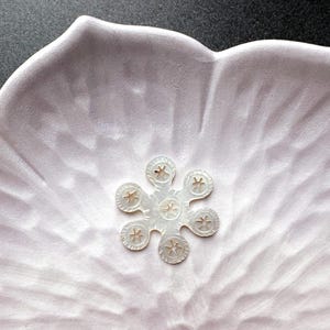 May include: A close-up of a light pink, petal-shaped ceramic dish with a white, flower-shaped embellishment in the center. The flower has six petals, each with a star-like design. The background is a dark gray.