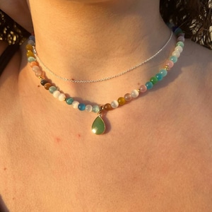 Può includere: Una collana a più strati con un ciondolo a forma di lacrima. La collana è realizzata con piccole perline colorate e una catena d'argento. Il ciondolo è di colore verde chiaro.