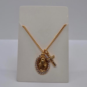 Può includere: Collana dorata con un ciondolo raffigurante una figura religiosa circondata da cristalli trasparenti e un ciondolo a forma di croce più piccolo. La collana è esposta su una carta bianca, che mette in risalto la sua delicata catena e il suo design intricato. Un gioiello a tema religioso.