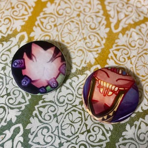 May include: Two circular badges with vibrant illustrations. One shows a smiling face with red lips, the other has a design with purple and green details. The badges are displayed on a patterned yellow and white surface.