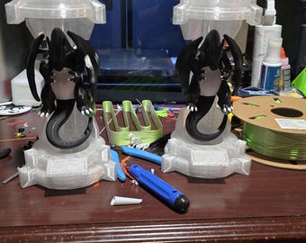 May include: Two black dragon figurines with outstretched wings, perched atop light gray pedestals. The dragons have detailed features and are set on a cluttered desk with 3D printing supplies, including a spool of green filament.