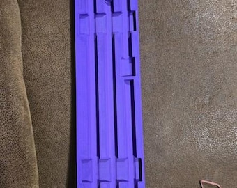 May include: A rectangular, purple 3D-printed object with a series of vertical and horizontal rectangular cutouts. The object is resting on a brown surface. The design appears to be a mold or a template.