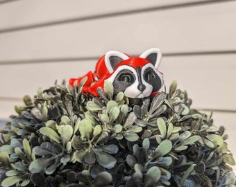 May include: A red, black, and white raccoon figurine rests atop a green and gray artificial topiary. The raccoon has a red body, white ears, and a black mask and nose. The topiary is a sphere shape with small, rounded leaves.