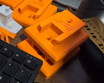 May include: Several bright orange, 3D-printed brackets with the "Milwaukee" logo are stacked on a wooden surface. A black keyboard and a small white 3D-printed object are also visible.