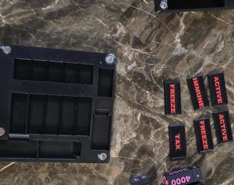 May include: Black game components including dice, trays, and labels with text like "FREEZE" and numbers. The dice have plus and minus symbols and numbers. The components are on a marble-patterned surface.