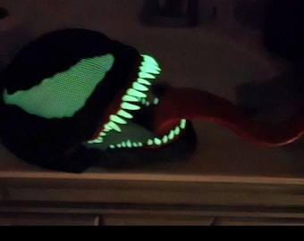 May include: A black mask with a glowing green eye and teeth, and a red tongue. The mask is designed to resemble a comic book character. The teeth and eye glow in the dark.