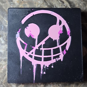 May include: Black square with a pink smiling face design. The pink design has a dripping paint effect. The item is likely a decorative piece or a small box.